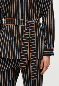 Μαύρο κοστούμι με κάθετες μπεζ και λευκές ρίγες, με ζώνη δεμένη που έχει πορτοκαλί περιγράμματα και παντελόνι με ταιριαστές ρίγες.