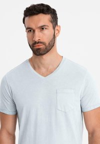 Světle modré tričko s krátkým rukávem a výstřihem do V, se zapínací kapsou na hrudi a měkkou texturou, s jednoduchými švy.
