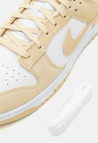 Gros plan d'une basket beige et blanche avec des lacets beiges et une paire supplémentaire de lacets blancs à côté sur fond blanc.