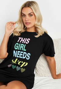 Svart bomulls-t-shirt med flerfärgad text: "DEN HÄR TJEJEN BEHÖVER sömn" och hjärtformade motiv i pastellfärger. Avslappnad passform, korta ärmar.