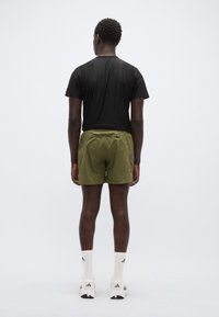 Ελιά πράσινα σορτς από ελαφρύ ύφασμα, συνδυασμένα με μαύρο t-shirt. Το μοντέλο φοράει λευκές αθλητικές κάλτσες και παπούτσια με ρίγες.