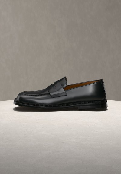 Μαύρο δερμάτινο μοκασίνι penny με ραφές, χαμηλό τακούνι και λείο γυαλιστερό φινίρισμα σε ουδέτερο φόντο.