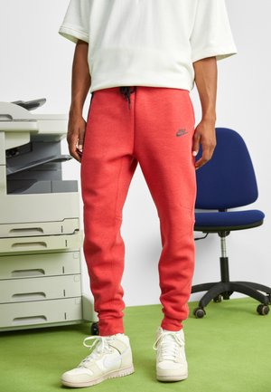Person trägt rote Nike-Jogginghosen und weiße Nike-Sneaker, steht auf grünem Teppich neben einem Bürostuhl und einem Drucker.