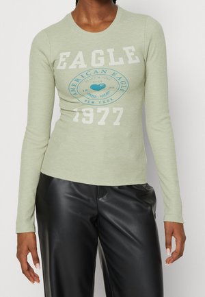 Person trägt hellgrünes Langarmshirt mit dem Text "EAGLE 1977 American Eagle New York" und schwarze Lederhose.