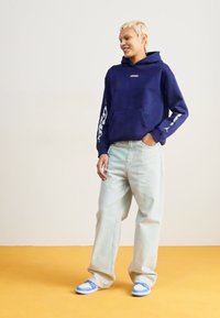 Camisola capuz azul marinho com bolso à frente e estampado gráfico branco, combinada com jeans largos azul claro e ténis com detalhes em azul, numa base amarela.