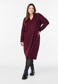 Burgundyröd stickad klänning med V-ringning, långa ärmar, lös passform och ribbad textur. Bärs med svarta ankelstövlar och strumpbyxor.