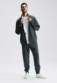 Szary dres sportowy składający się z zapinanej na zamek kurtki i pasujących spodni, wykonany z miękkiego materiału z prążkowanymi detalami. Całość dopełniają białe sneakersy.