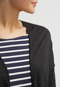 Sort let cardigan med åben front, lagt over en navy og hvid striped top. Blød tekstur, afslappet design og løs pasform.