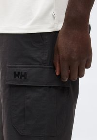 Gros plan sur une main à la peau foncée reposant près d'une poche cargo noire avec un logo "HH" noir sur un pantalon foncé et un ourlet de chemise blanc.