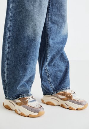 Personne portant un jean bleu ample et des baskets épaisses avec des détails beiges, blancs et dorés scintillants, debout sur un sol blanc.