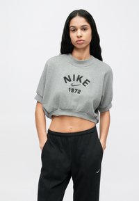 Γκρι cropped φούτερ με ρελιασμένα μανίκια, με μαύρο λογότυπο «NIKE» και «1972» από κάτω. Συνδυάζεται με μαύρο παντελόνι φόρμας.