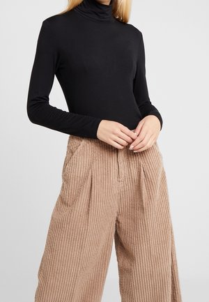 Personne portant un haut noir à col roulé à manches longues et un pantalon beige en velours côtelé à taille haute et jambes larges, les mains jointes devant.