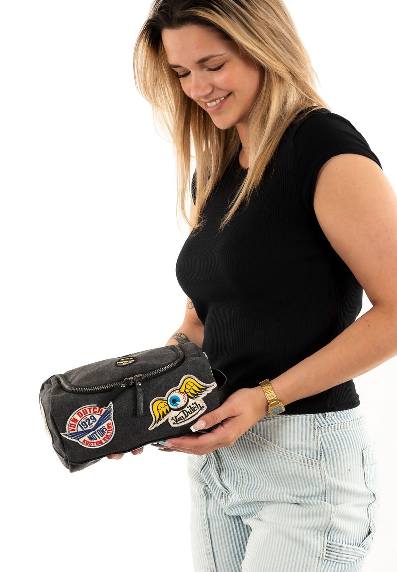 Borsa in tessuto grigia con chiusura a zip, caratterizzata da toppe colorate, inclusa un'occhio blu con ali e una toppa con logo. Forma compatta e rettangolare.