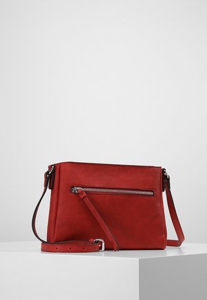 Sac bandoulière rectangulaire rouge avec sangle ajustable et poche avant zippée horizontale, posé sur une surface blanche contre un fond uni.