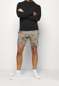 Černá mikina s kulatým výstřihem, spárovaná s khaki šortkami s jemným vzorem palmového listu. Model má na sobě bílé tenisky.