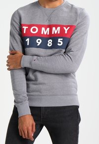 Sudadera gris con rayas horizontales rojas y marinas, presenta "TOMMY" y "1985" impresos en blanco, puños acanalados y cuello redondo.