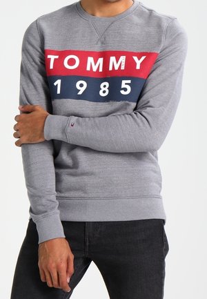 Sweat à capuche gris avec des rayures horizontales rouges et marines, affiche "TOMMY" et "1985" imprimés en blanc, avec des poignets côtelés et un col rond.