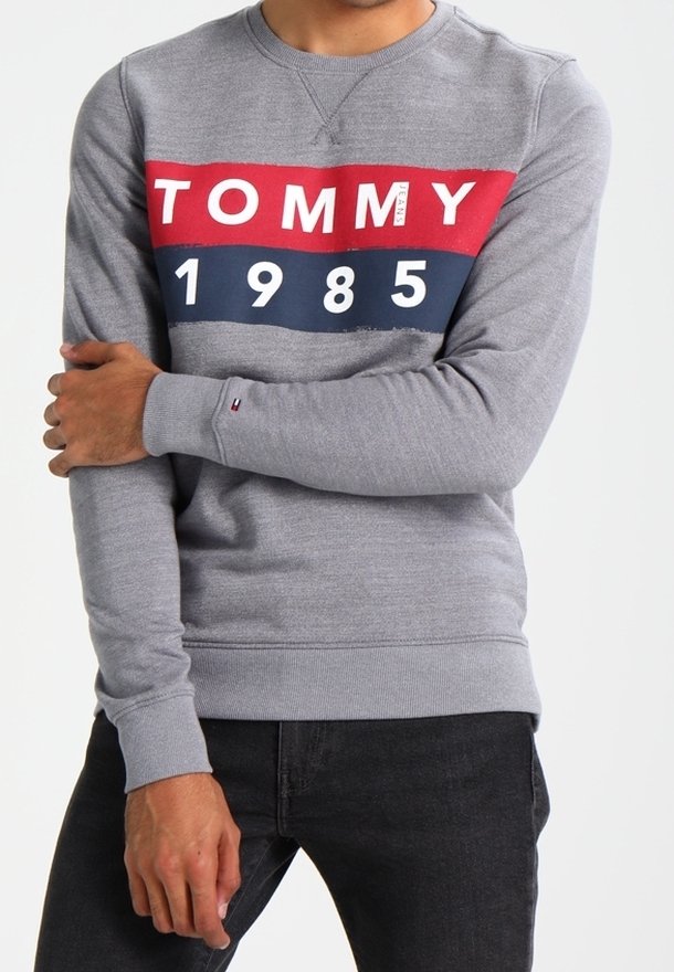 Sudadera gris con rayas horizontales rojas y marinas, presenta "TOMMY" y "1985" impresos en blanco, puños acanalados y cuello redondo.