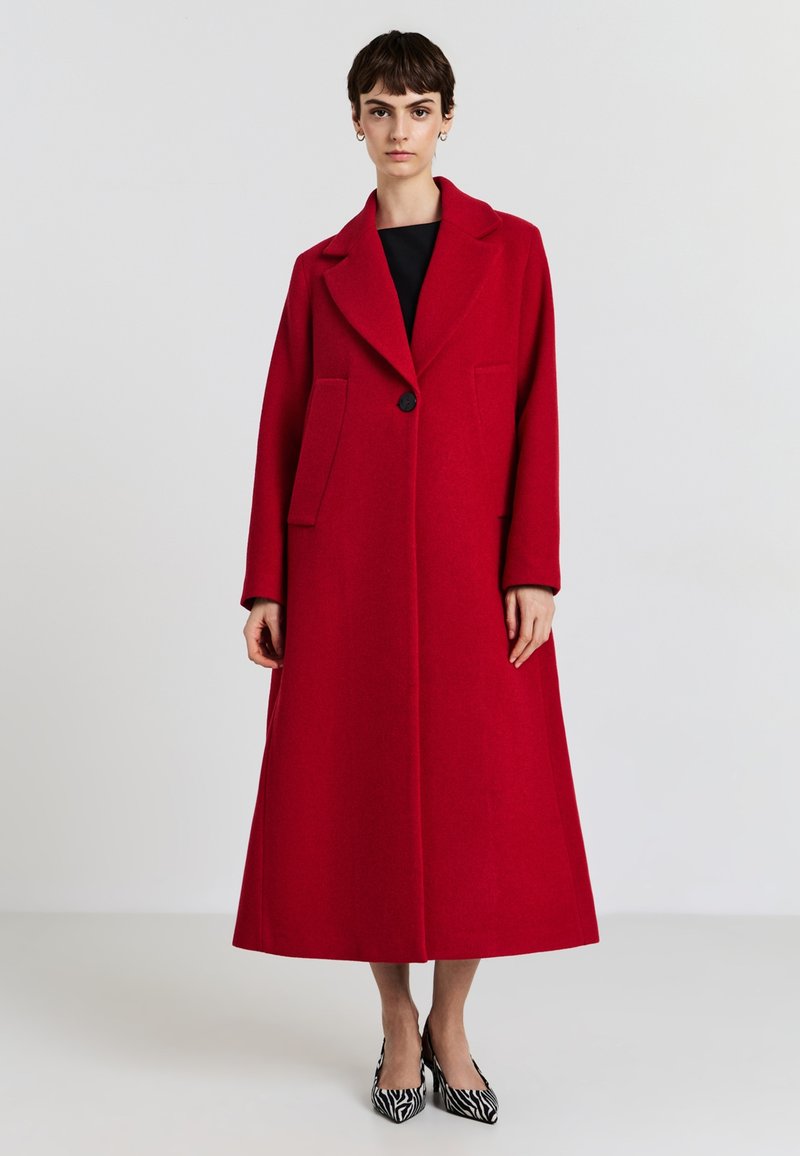 Roter Wollmantel mit Reverskragen, Ein-Knopf-Verschluss und zwei Fronttaschen. Er zeichnet sich durch eine lange, A-Linien-Form und eine glatte Textur aus.