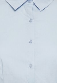 Chemise bleu clair à col avec surpiqûres visibles et quatre boutons transparents sur la patte de boutonnage avant.