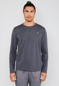 Chemise à manches longues en tissu gris foncé, col rond, arborant un petit logo blanc sur la poitrine. Texture douce avec une coupe décontractée.