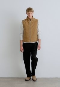 Rjava semšena telovnik preko belega rebrastega puloverja in črnih hlač. Telovnik ima zadrgo in je kombiniran z bež gležnjaki.