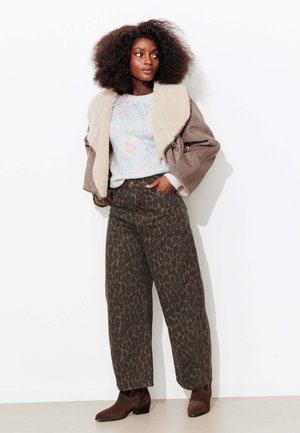 Femme aux cheveux bouclés portant une veste en peau lainée beige, un pull fleuri, un pantalon à imprimé léopard et des bottines marron, debout contre un mur blanc.