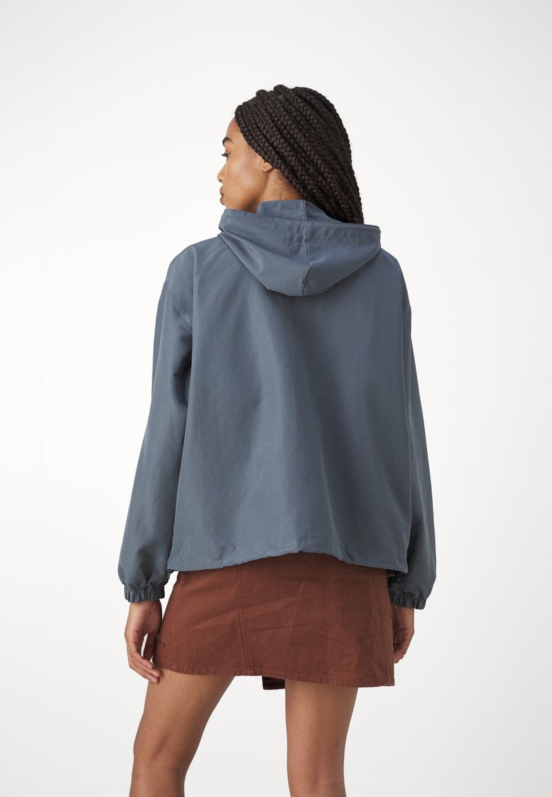 Blaues Kapuzenjackett mit elastischen Bündchen und lässiger Passform, kombiniert mit einem braunen Cordrock. Glatte Textur und minimalistisches Design.
