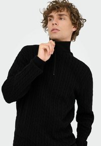 Junger Mann mit lockigem Haar, der den Reißverschluss auf einem schwarzen Strickpullover mit hohem Kragen anpasst, vor einfachem Hintergrund.