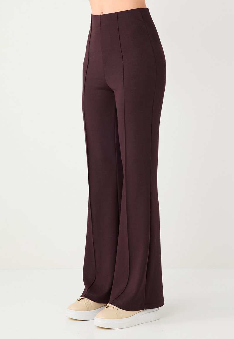 Pantalon évasé taille haute de couleur marron foncé avec des coutures verticales, fabriqué en tissu doux. Associé à des baskets compensées de couleur claire.