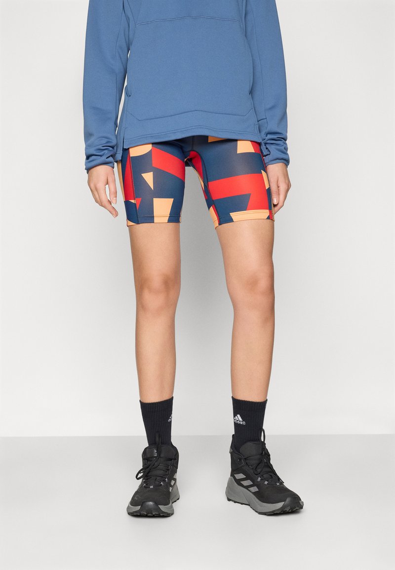 Shorts sportivi colorati presentano motivi geometrici in rosso, arancione e blu navy. Abbinati a una maglietta a maniche lunghe blu e scarpe nere.