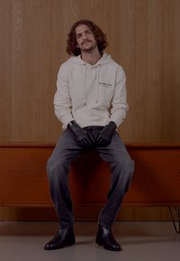 Balta kapuce ar auklītēm, pelēki džinsu bikses un melni cimdi atrodas uz koka soliņa. Tumšie potīšu zābaki pabeidz apģērbu.