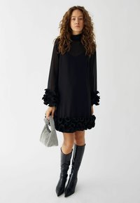 Črna obleka z prosojnimi dolgimi rokavi, z nabranimi črnimi detajli na manšetah in robu, v kombinaciji z visokimi črnimi špičastimi škornji do kolen in torbico v houndstooth vzoru.