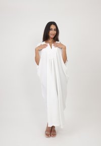 Kaftan blanc avec un décolleté en V profond et de larges manches. Tissu fluide avec une texture légère, design long. Porté sur des sandales transparentes à talons.