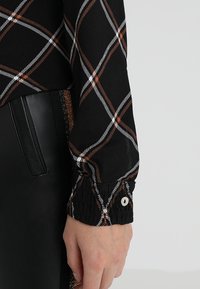 Blusa a scacchi nera con motivi a diamante bianchi e ruggine, con maniche testurizzate e polsini con bottoni. Pantaloni in pelle neri visibili.
