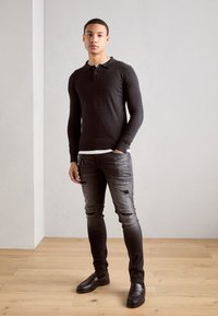 Mørk langærmet polo med tre-knaps lukning, parret med slidte gr�å jeans og sorte slip-on sko. Enkel design, strikket tekstur.