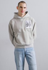 Jasnoszara bluza z kapturem z kieszenią z przodu, z naszywką z nadrukiem zdjęcia. W połączeniu z jasnoniebieskimi dżinsami, o luźnym kroju.