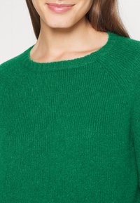 Femme portant un pull tricoté vert vif avec un col rond et une texture douce, souriant légèrement, longs cheveux bruns visibles.