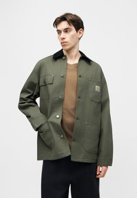 Giovane uomo con giacca verde oliva abbottonata e colletto nero sopra una camicia marrone, in piedi con una mano in tasca, che guarda a sinistra.