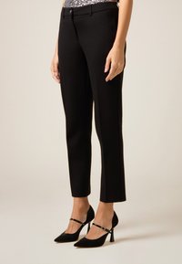 Zwarte, op maat gemaakte broek met een gladde textuur, een slim-fit pasvorm en een subtiele voorplooi. Gecombineerd met zwarte pumps met spitse neus en enkelriempjes.