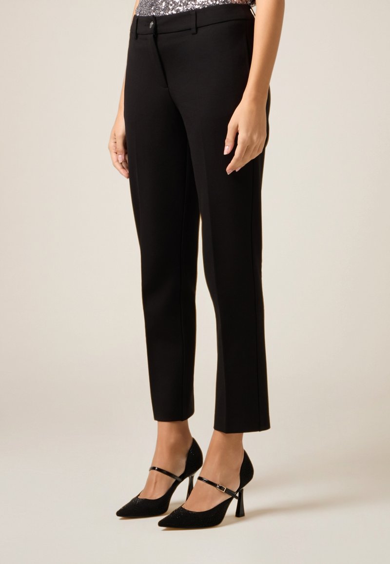 Zwarte, op maat gemaakte broek met een gladde textuur, een slim-fit pasvorm en een subtiele voorplooi. Gecombineerd met zwarte pumps met spitse neus en enkelriempjes.