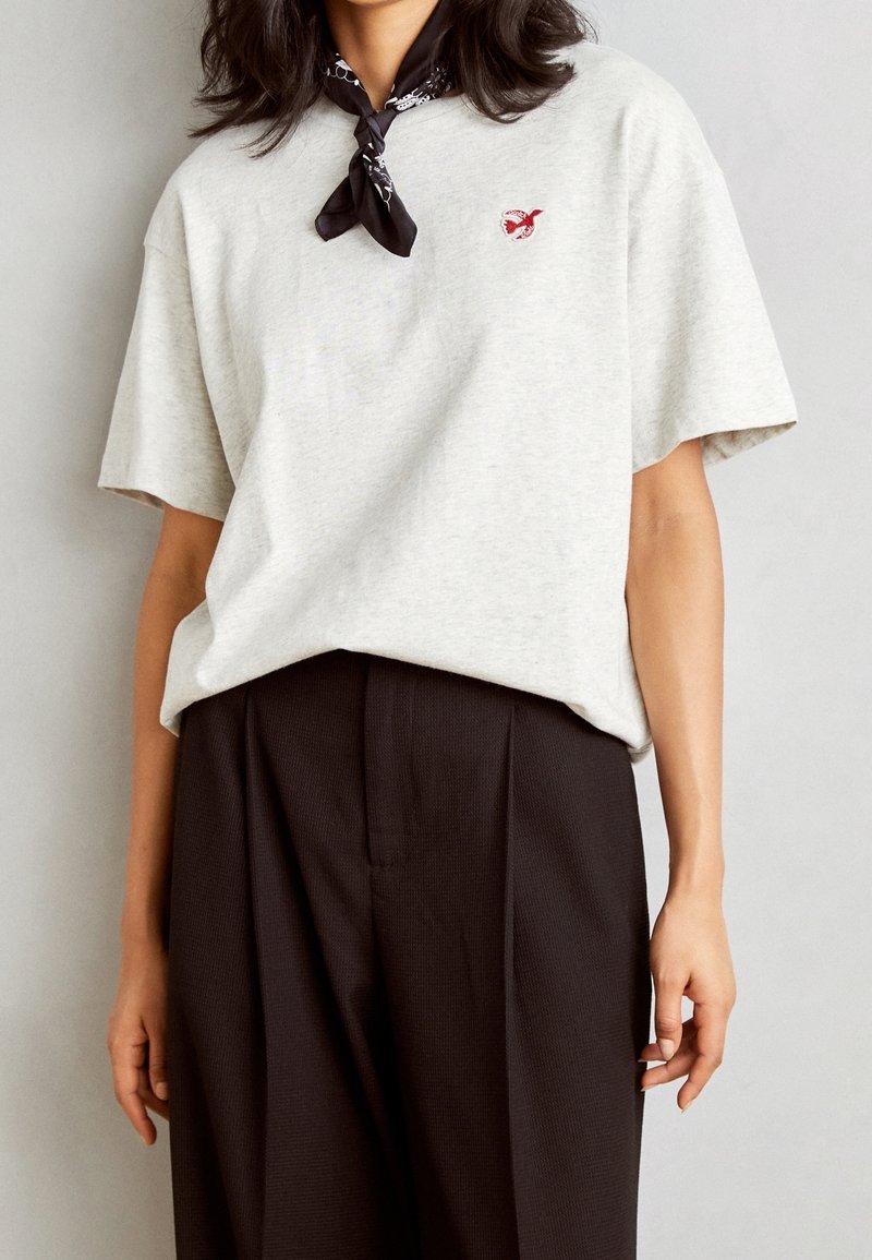 Jasnoszary, oversize'owy T-shirt wykonany z bawełny; z małym czerwonym haftowanym logo oraz czarnym wzorzystym szalem na szyi. W zestawie z ciemnymi spodniami w kant.