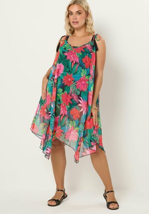 Kvinde iført ærmeløs grøn kjole med pink og rød tropisk blomstermønster, sorte sandaler og guldbøjle, stående mod hvid baggrund.