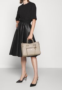 Miękka beżowa skórzana torebka z przednią kieszenią i złotymi okuciami, trzymana przez osobę w czarnej bluzce i czarnej skórzanej spódnicy.