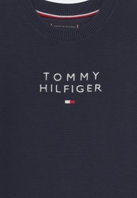 Granatowy sweter z dzianiny z okrągłym dekoltem, teksturowaną tkaniną i haftowanym logo "TOMMY HILFIGER" z czerwono-białym akcentem poniżej.