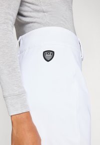 Λευκό αθλητικό παντελόνι με ομαλή υφή, που έχει μαύρο λογότυπο EA7 στη μέση και ανοιχτό γκρι μακρυμάνικη μπλούζα.