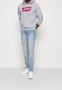 Grå huvtröja med vita dragkedjor och röd Levi's-logga, tillsammans med ljusblå jeans och vita sneakers. Enkel, avslappnad outfit.