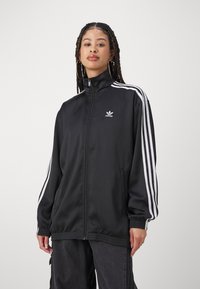 Chaleco negro con cremallera hecho de una tela suave, con franjas blancas en las mangas. Presenta un pequeño logo de Adidas en el pecho y puños elásticos.