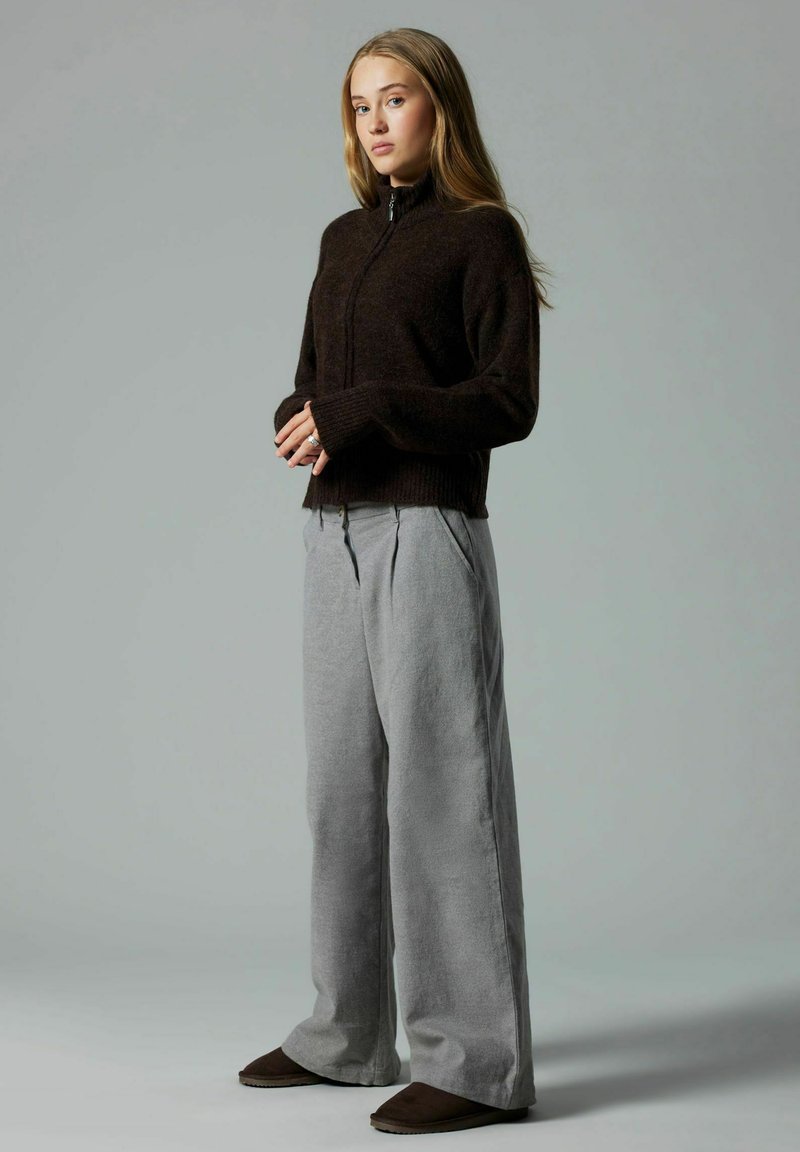 Jeune femme debout, les mains jointes, vêtue d'un pull zippé foncé, d'un pantalon large gris clair et de chaussures foncées, sur un fond gris uni.