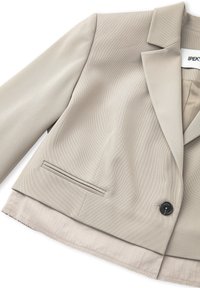 Blazer beige su misura con risvolto a soffietto, chiusura con un bottone nero singolo, texture elegante e una tasca sul petto. Fodera interna visibile.